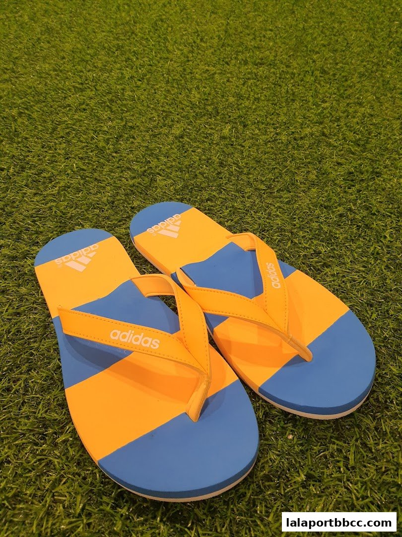 A pair of blue and orange Adidas flip-flops displayed on a green grassy surface.