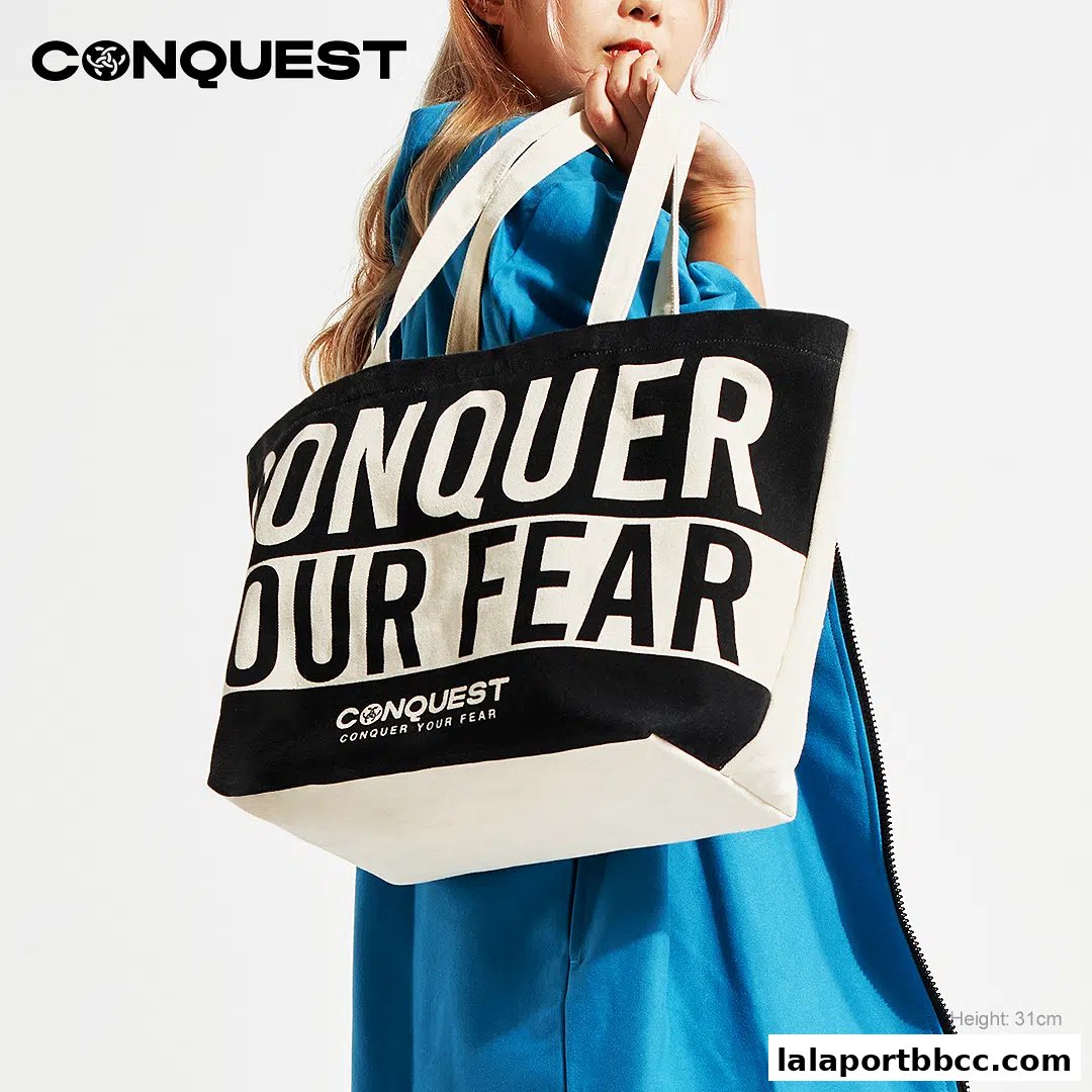 A person holds a large black and white tote bag with bold text that says 'CONQUER OUR FEAR'.