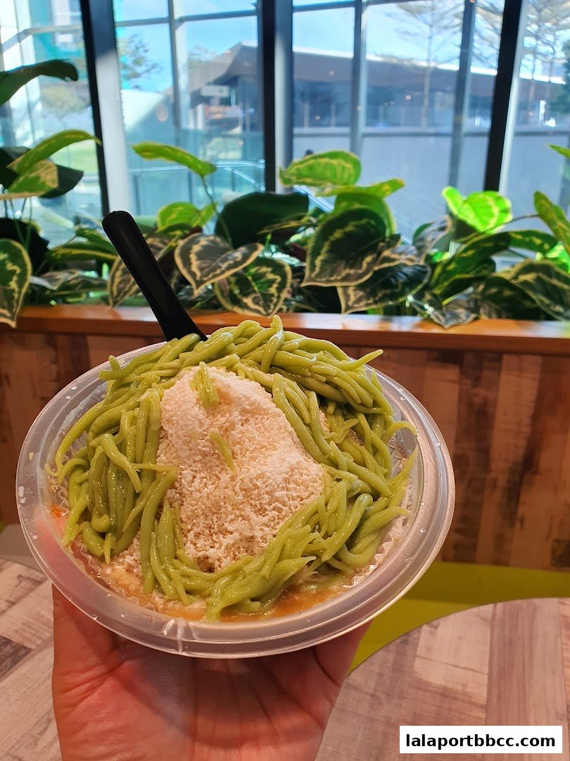 A hand holds a clear plastic bowl filled with green cendol dessert topped with shaved ice.