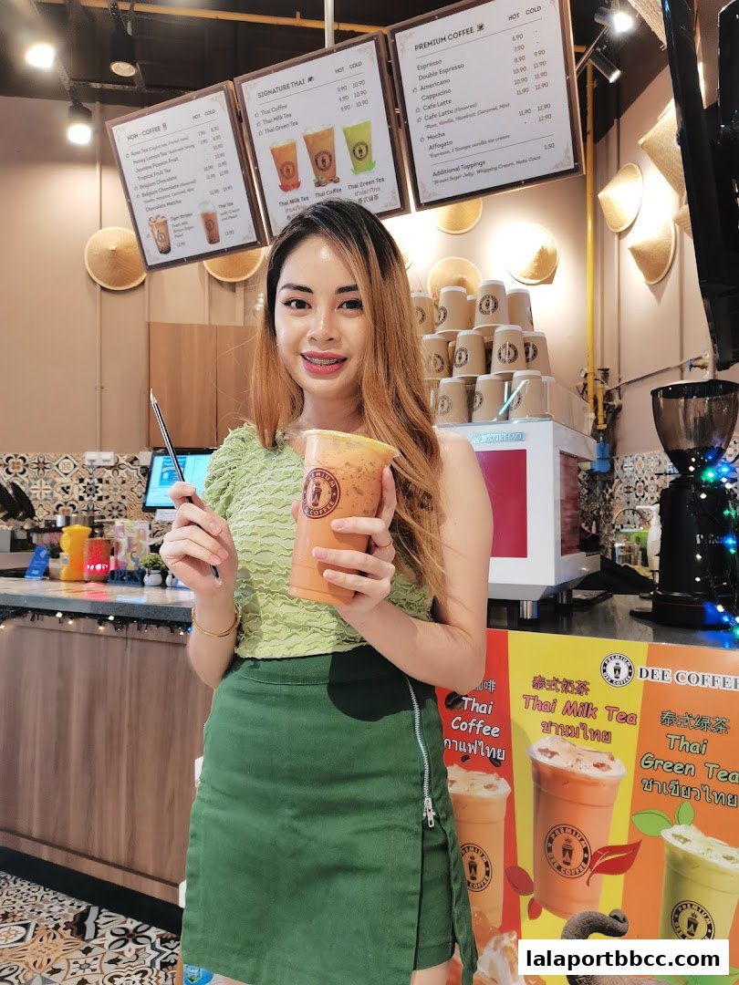 A woman holds a Thai milk tea in front of a menu board at Dee Coffee Malaysia.