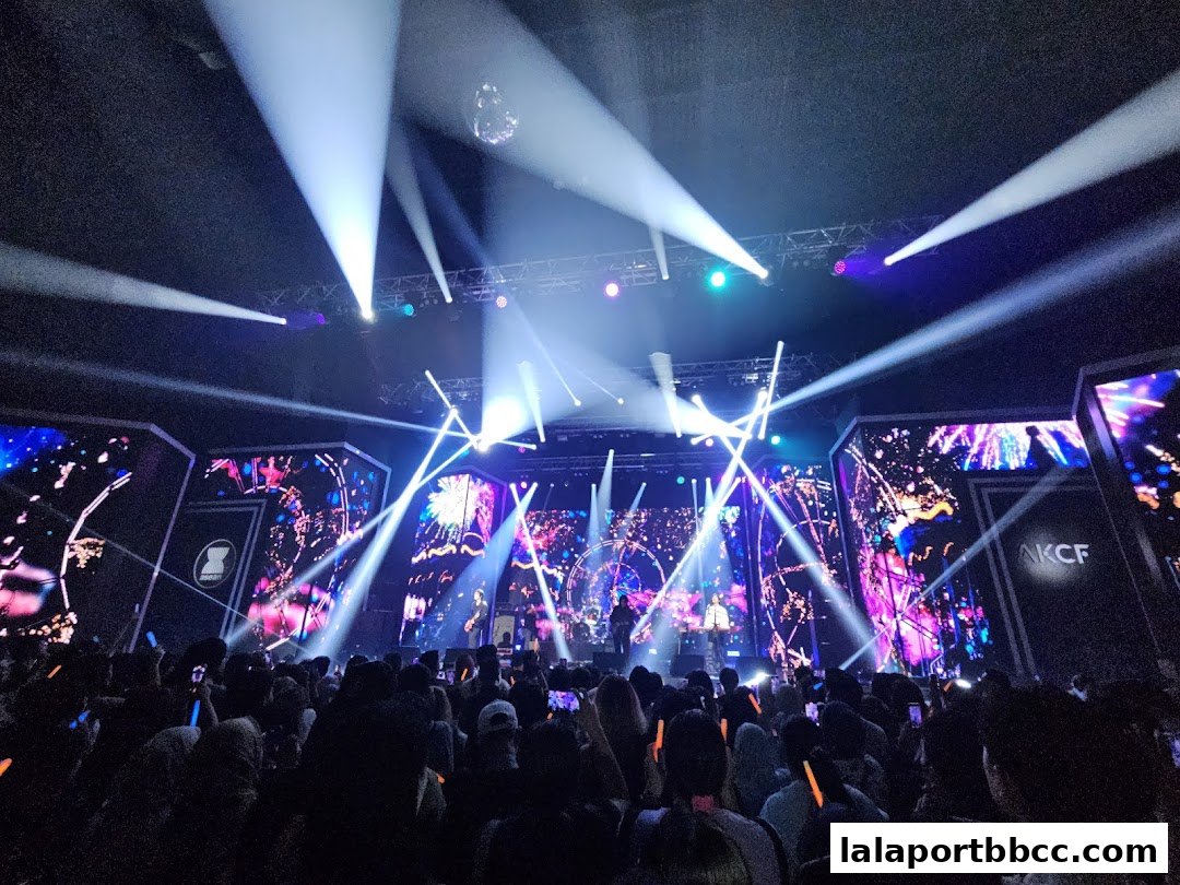 A concert stage with bright lights and a large audience holding glowing sticks.