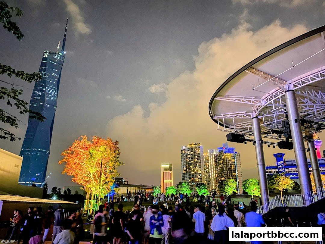 Night view of a rooftop garden with a crowd, a tall skyscraper, and illuminated trees.