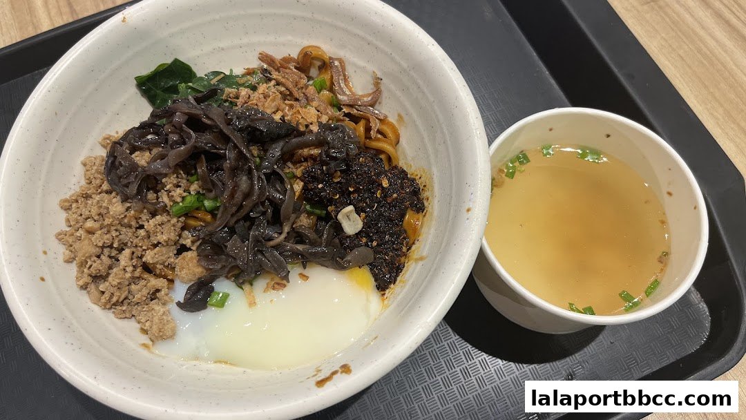 A bowl of chili pan mee with minced meat, mushrooms, and a soft-boiled egg, served with a side of soup.