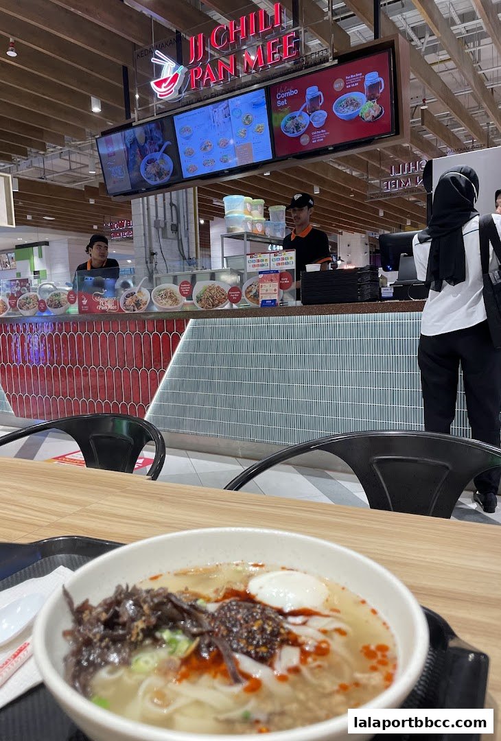 A close-up of a bowl of pan mee with toppings, with the restaurant counter and staff in the background.
