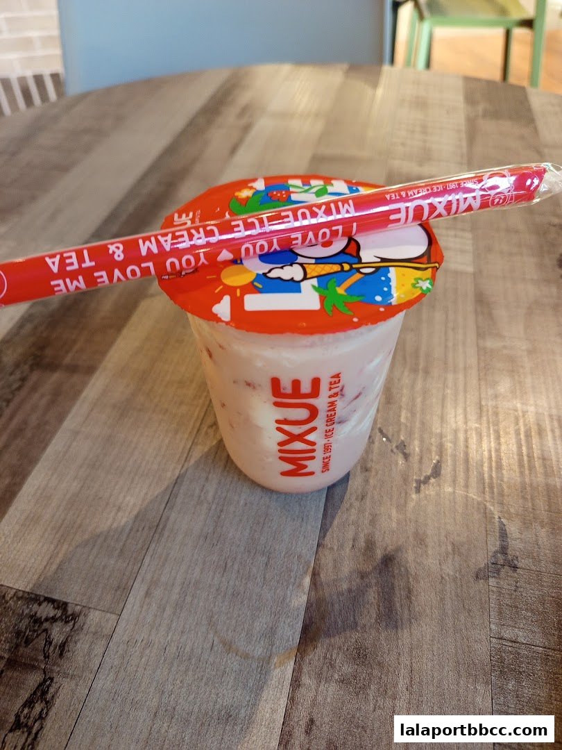 A cup of pink strawberry ice cream and tea with a red straw on a wooden table.