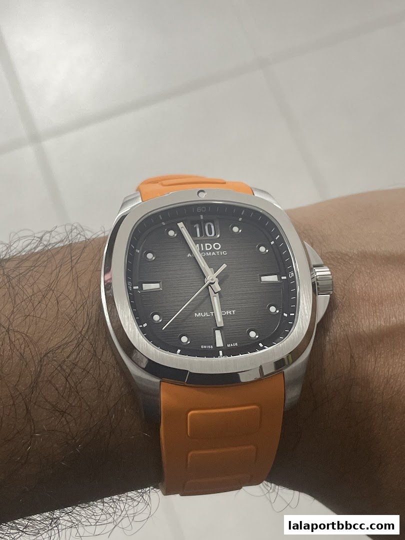 A close-up shot of a Mido automatic watch with a grey dial and an orange rubber strap on a wrist.