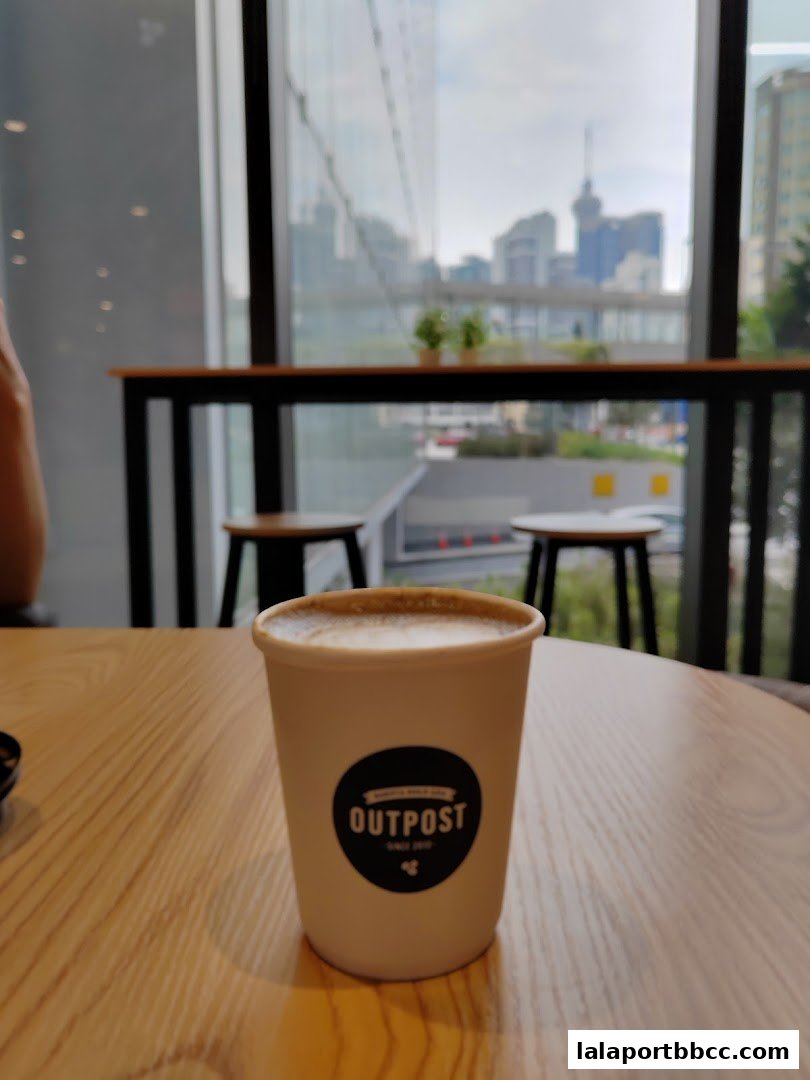 A cup of coffee with the 'Outpost' logo sits on a wooden table, with a city view through the window.