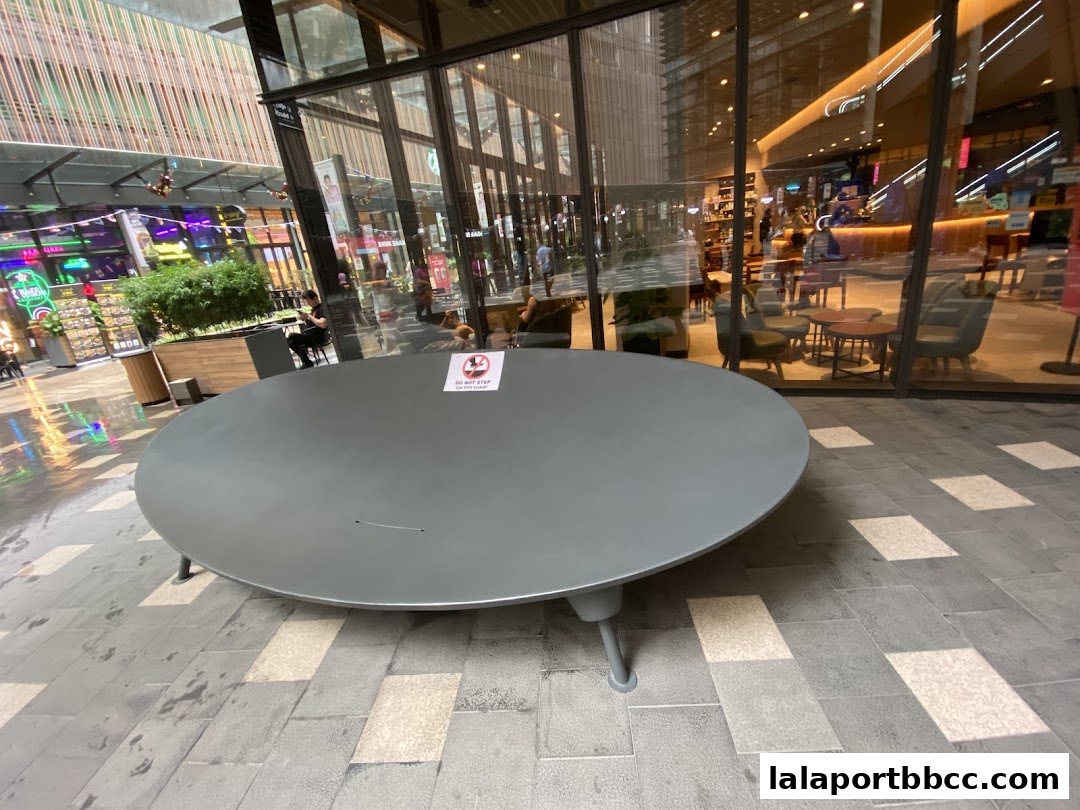 A large, round table sits in an outdoor plaza with shops reflected in the background.
