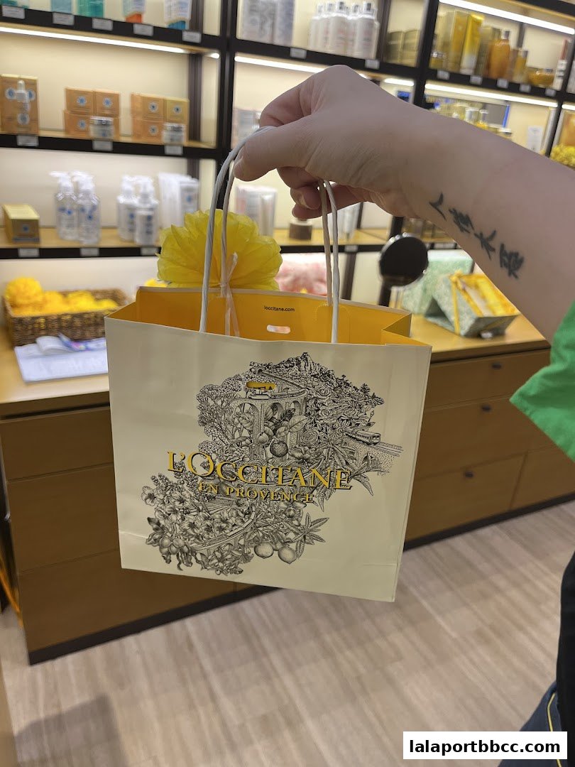 A hand holds a L'Occitane shopping bag with products displayed in the background.