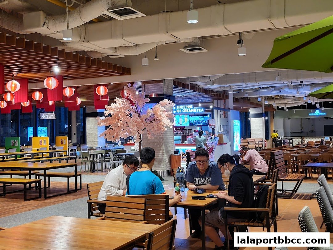 People dining at tables in a brightly lit food court with decorative lanterns and a cherry blossom tree.