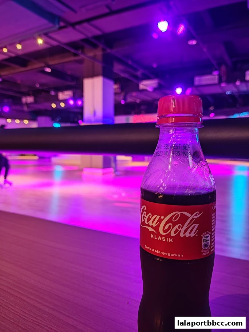 A bottle of Coca-Cola sits on a wooden surface with a roller skating rink in the background.