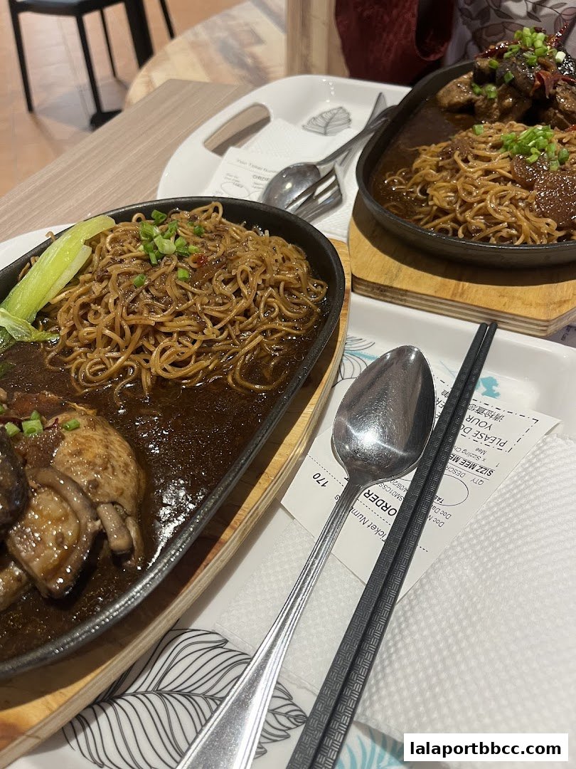 Two sizzling noodle dishes served in cast iron pans with chopsticks and spoons.