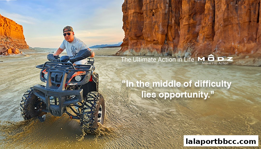 A man rides an ATV through water with rocky cliffs in the background.