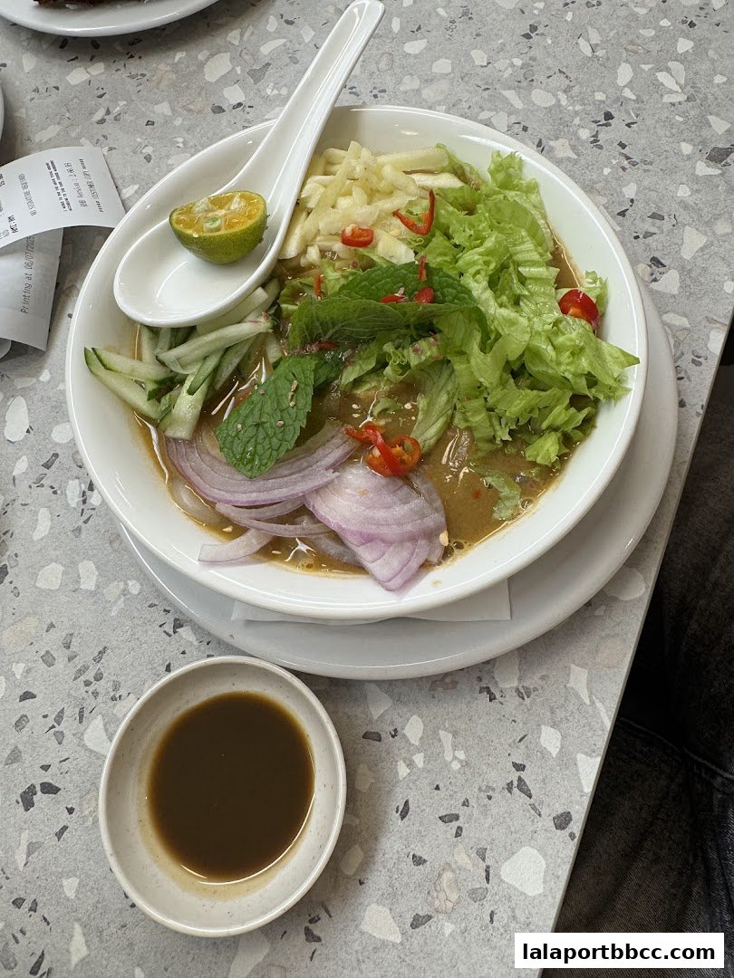 A bowl of Laksa with fresh ingredients and a side of dipping sauce.