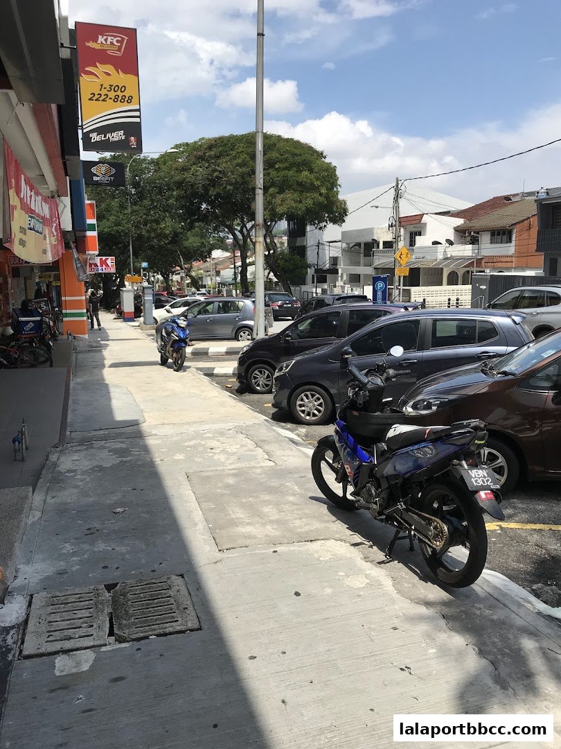 A street view showing parked cars and motorcycles in front of shops.
