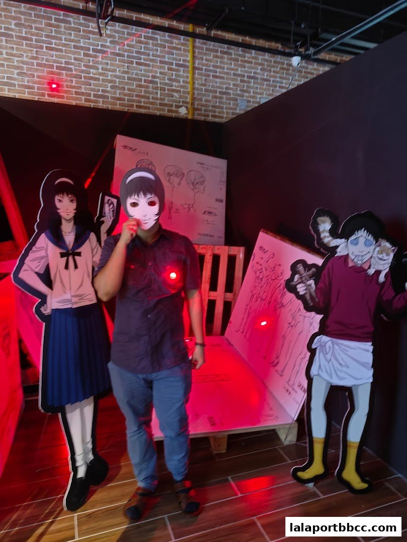 A person holds a mask in front of their face, standing between Junji Ito character cutouts.
