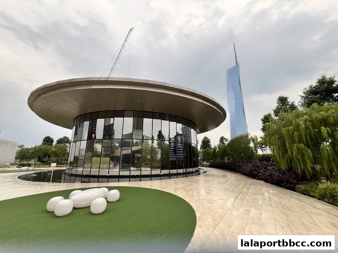 Modern circular building with glass walls on a rooftop garden with a distinctive skyscraper in the background.