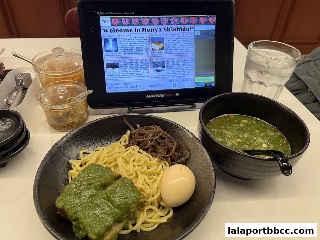 A bowl of ramen with noodles, meat, egg, and broth, next to a tablet and condiments.