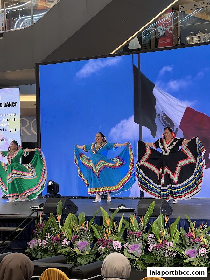Three women in traditional Mexican dresses perform a folk dance on a stage with a Mexican flag backdrop.