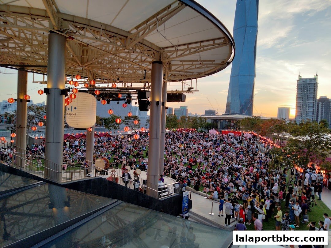 A large crowd gathers on a grassy area under a canopy decorated with lanterns.
