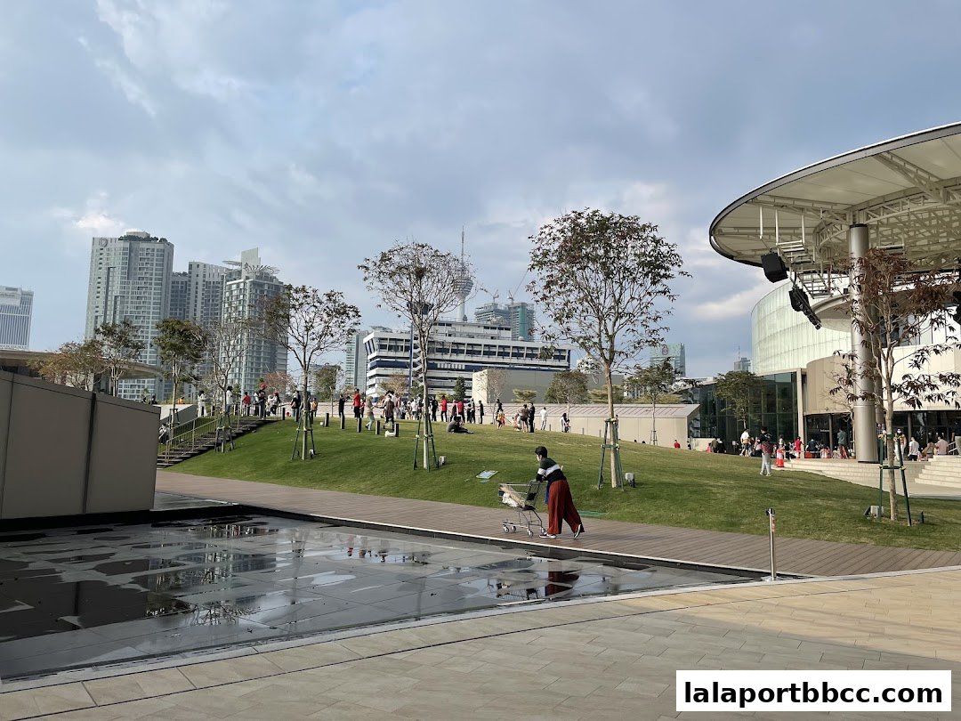 People enjoying a sunny day in a park with modern buildings and an outdoor stage.