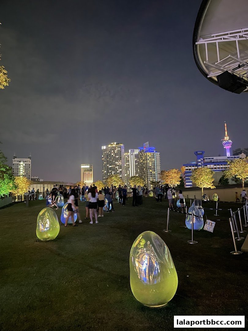 People gather on a grassy rooftop at night, surrounded by illuminated art installations and city skyline.