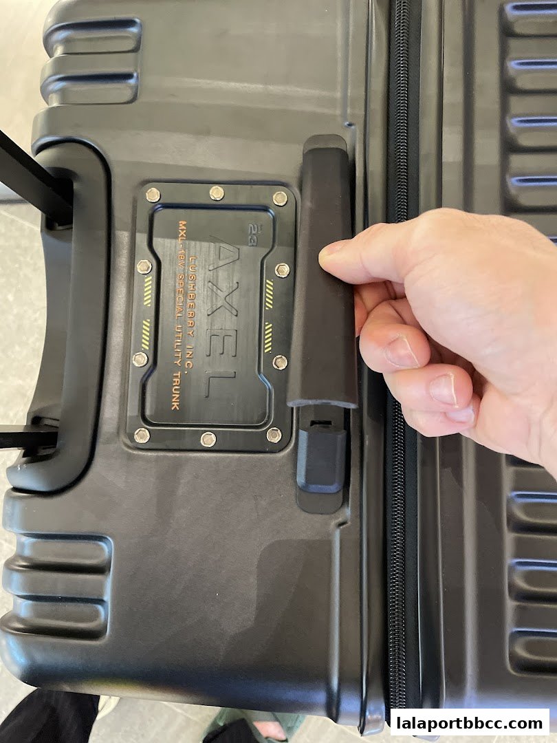 A hand is shown operating the handle of a rugged, dark grey suitcase with 'AXEL' branding.
