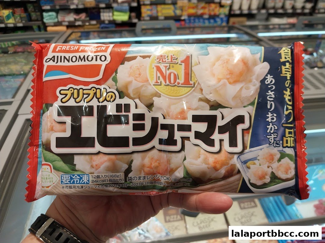 A hand holds a package of Ajinomoto frozen shrimp shumai in a grocery store freezer aisle.