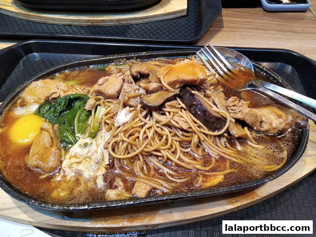 A sizzling claypot dish with noodles, meat, mushrooms, and a fried egg.