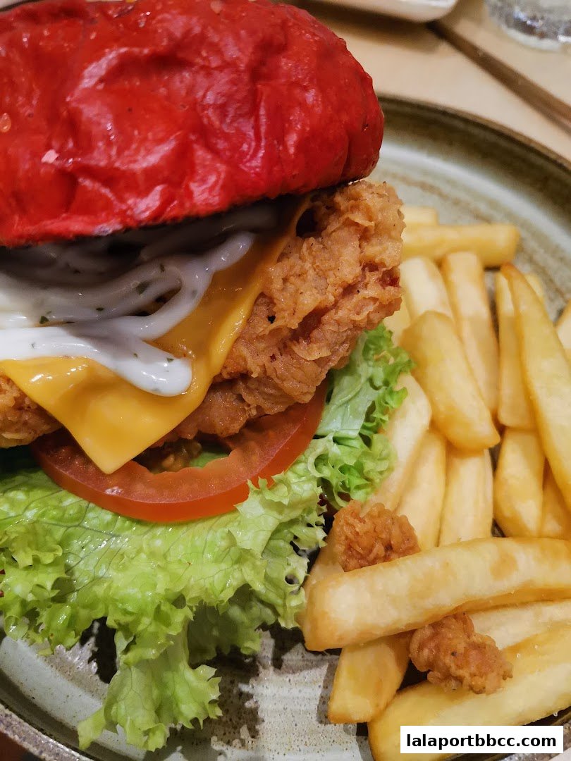 A close-up shot of a gourmet burger with a red bun, crispy chicken, cheese, and fries.