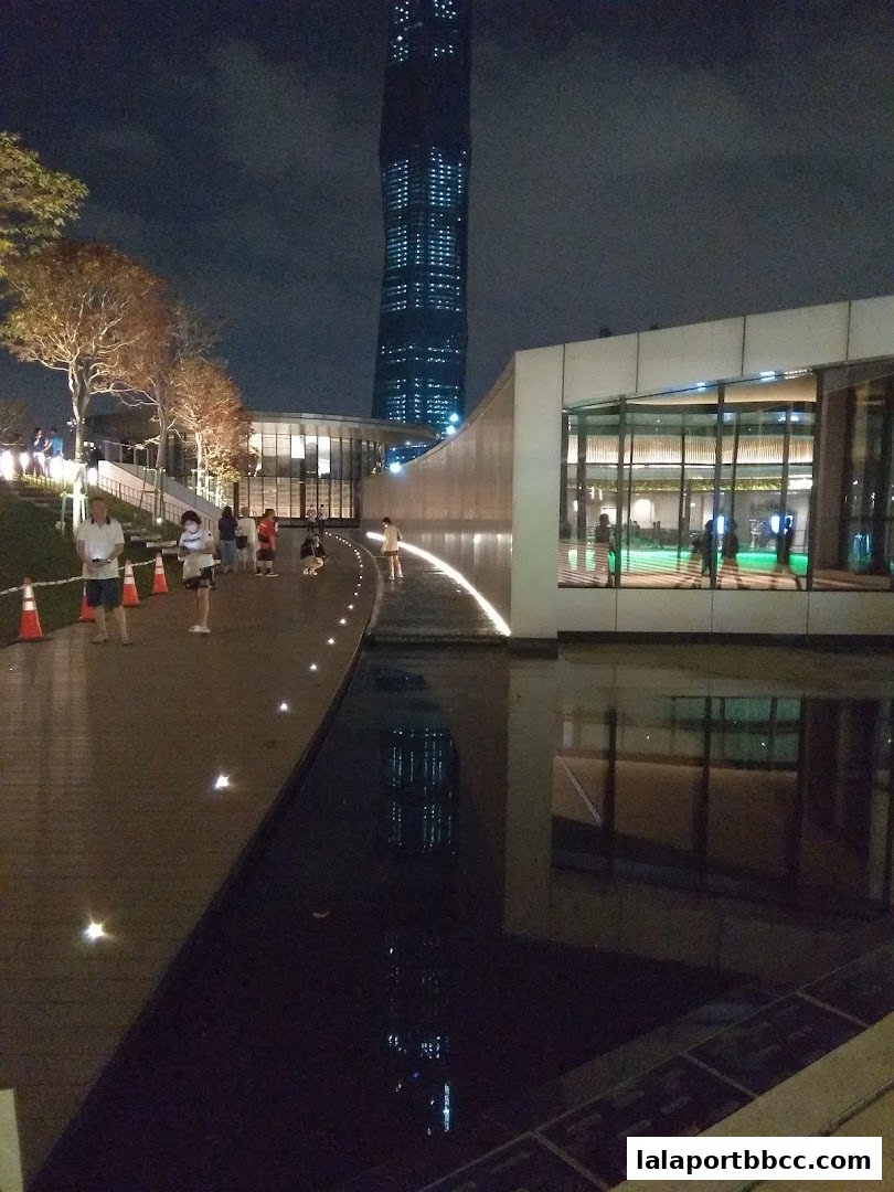 A modern building with large windows and a reflective pool at night, with a skyscraper in the background.