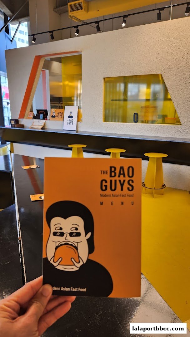 A hand holds a menu for 'The Bao Guys' featuring a cartoon character eating a bao.