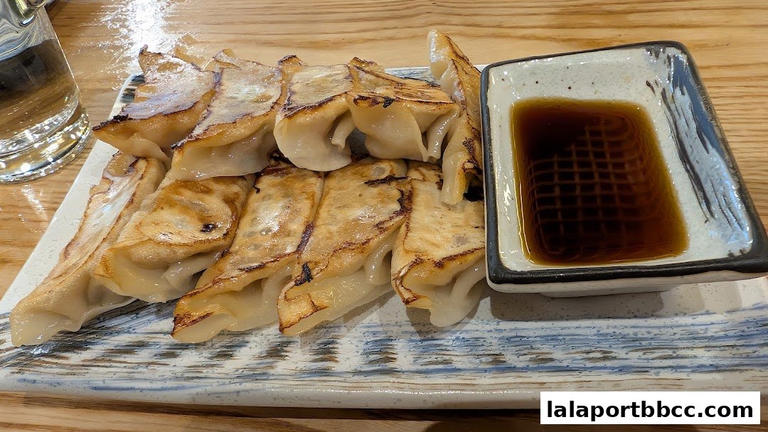 A plate of golden-brown pan-fried gyoza with a side of soy sauce and a glass of water.