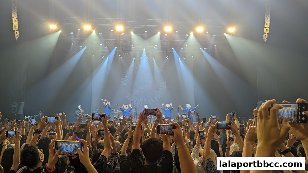 A band performs on stage with a crowd holding up phones to record.