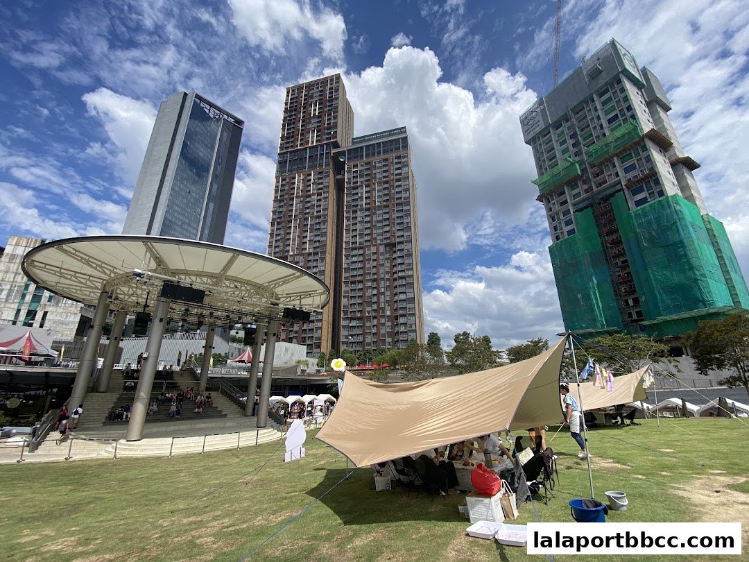Outdoor event with tents and people gathered under a canopy with skyscrapers in the background.