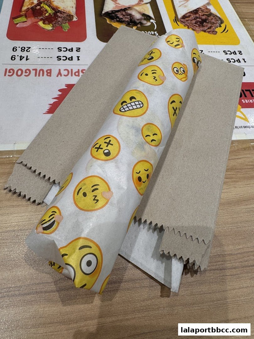 A kebab wrapped in paper with various emojis, placed on a table with menu items visible.