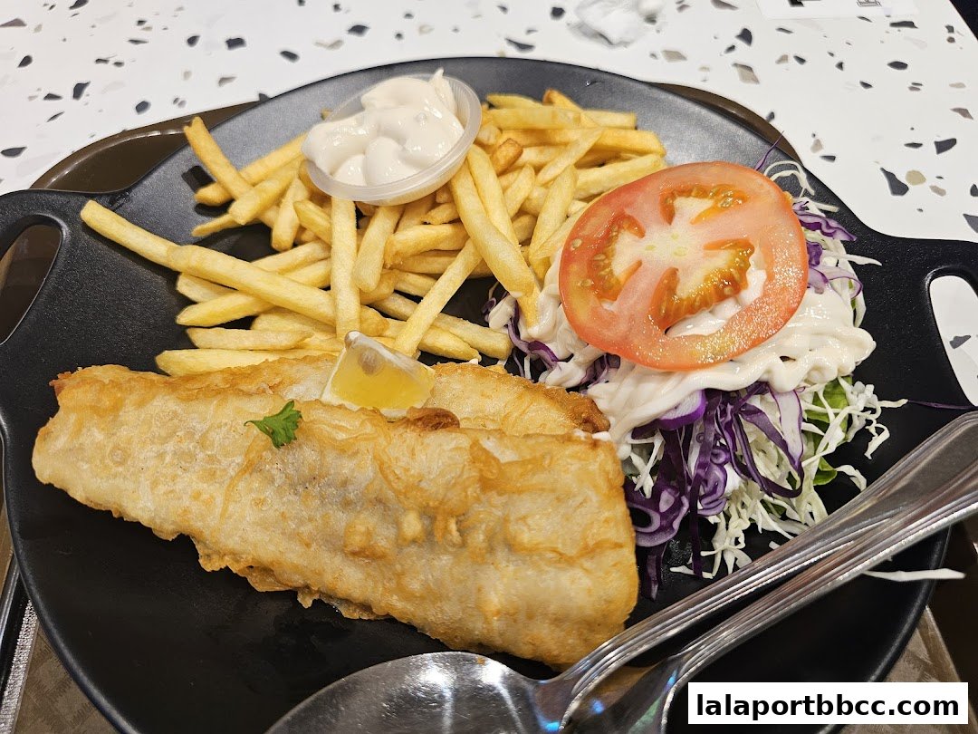 A plate of crispy fish and chips with coleslaw and a slice of tomato.