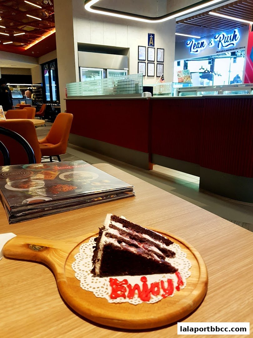 A slice of chocolate cake with red frosting on a wooden board, with menus and a cafe interior in the background.