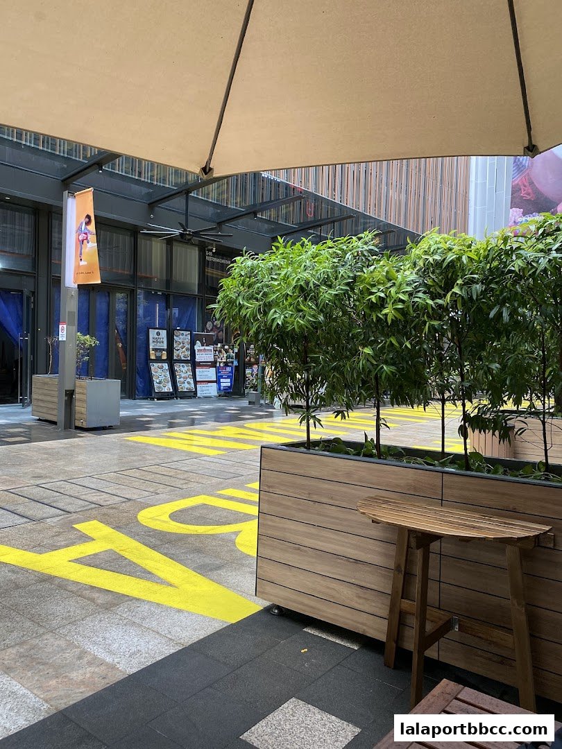 An outdoor seating area with plants and tables, leading to shops with signage.