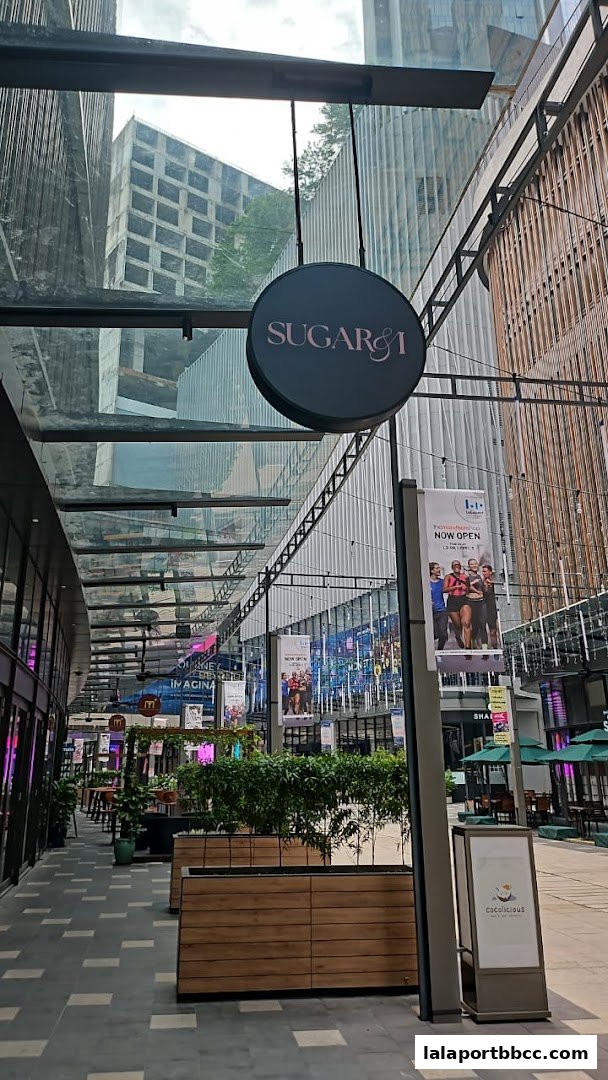 A circular sign for Sugar & I hangs above a walkway in a modern shopping mall.