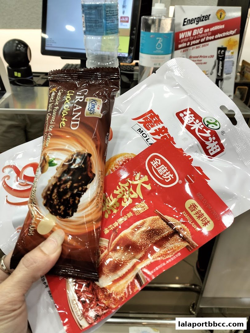 A hand holds various packaged snacks in front of a store counter with a computer monitor and products.