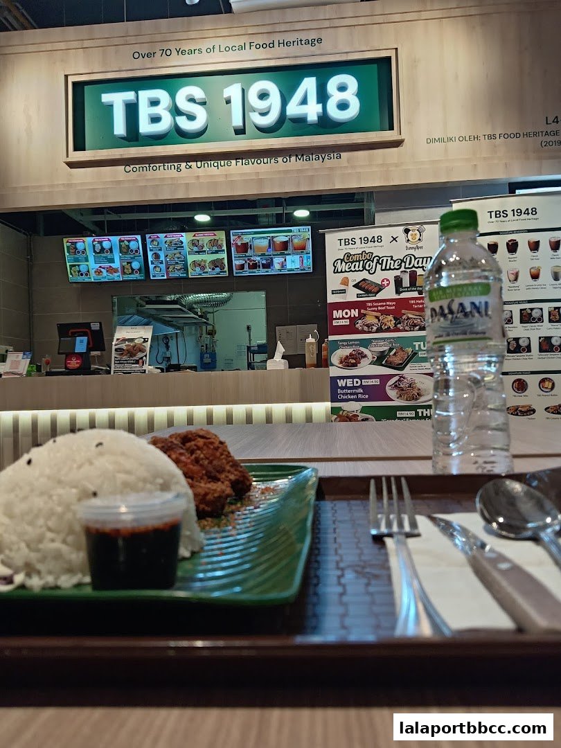 A plate of rice with fried chicken and dipping sauce, with a bottle of water and the TBS 1948 shop front in the background.