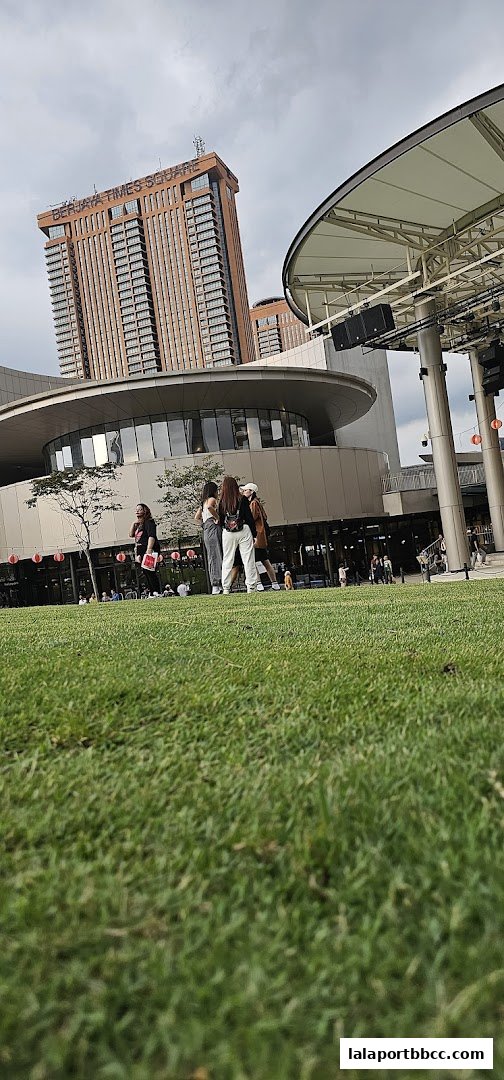 People gather on a grassy area in front of a modern building complex with a tall tower.