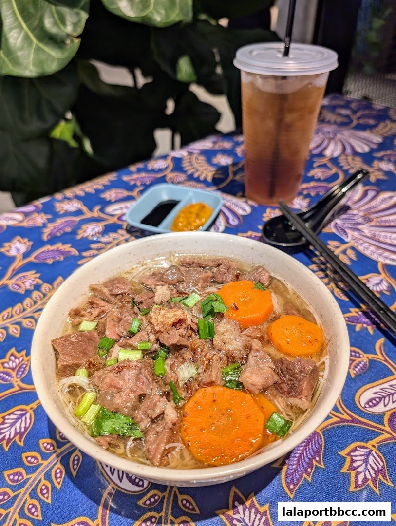 A bowl of beef noodle soup with carrots and a side of dipping sauce and iced tea.