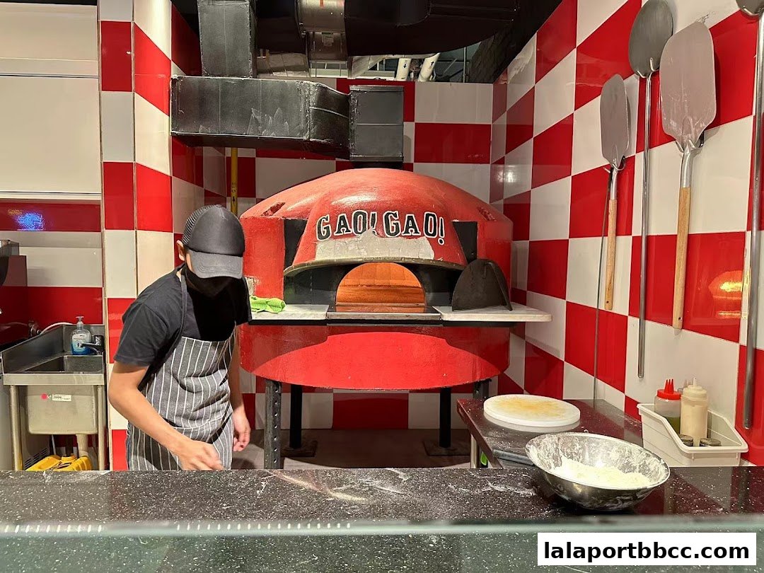 A chef prepares dough near a traditional red pizza oven with pizza peels on the wall.