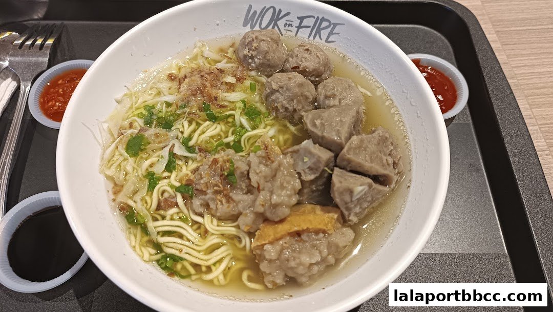 A bowl of noodles with meatballs and broth, served with chili sauce and soy sauce.