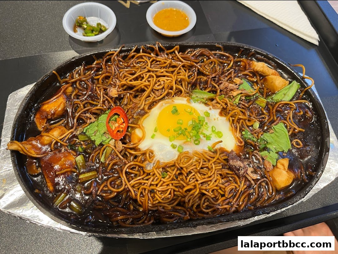 A sizzling plate of noodles with a fried egg, chili, and greens, served with dipping sauces.