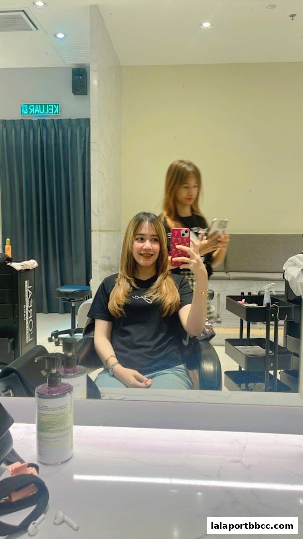 A woman takes a selfie in a salon chair with a stylist in the background.