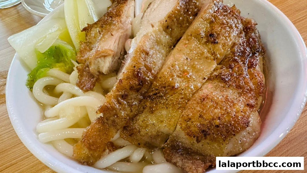 A close-up of a bowl of udon noodles topped with sliced, crispy roasted chicken and greens.