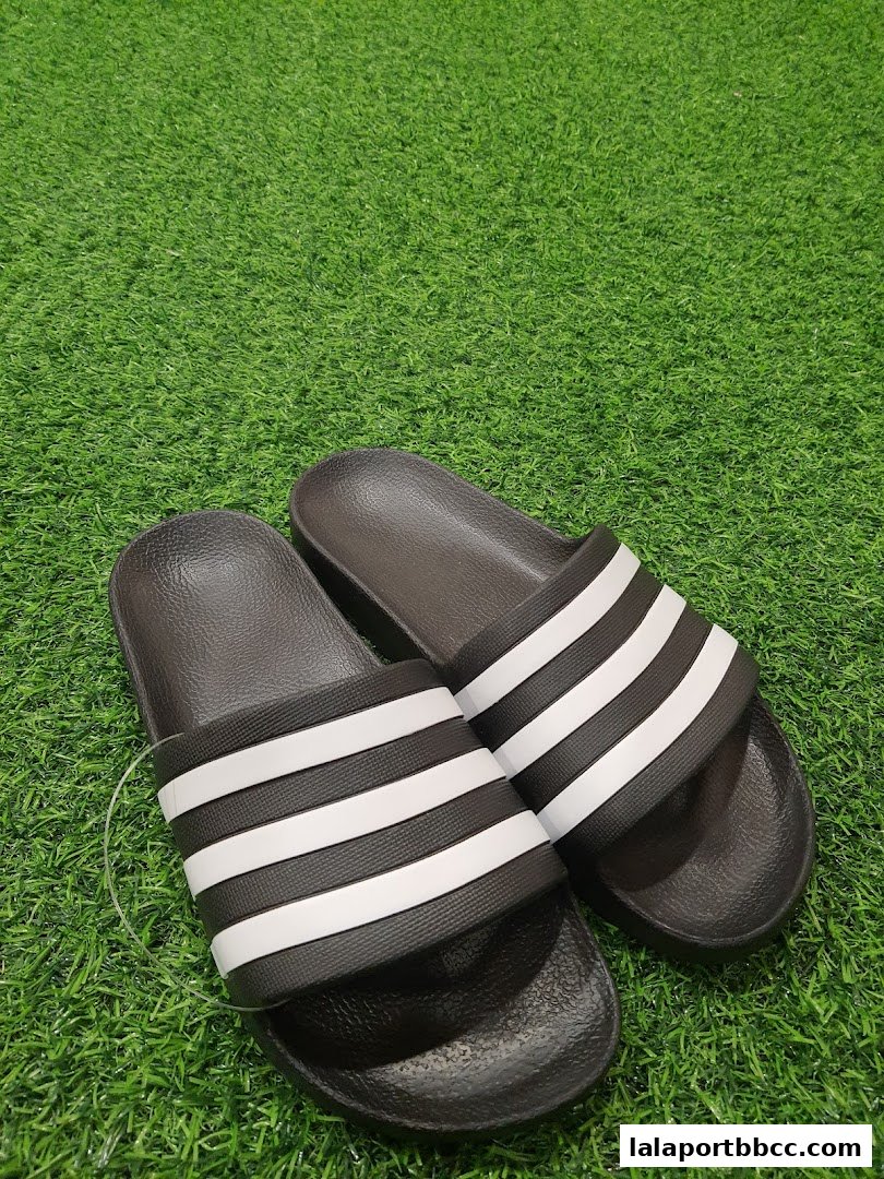 A pair of black Adidas slides with white stripes on a green grassy background.
